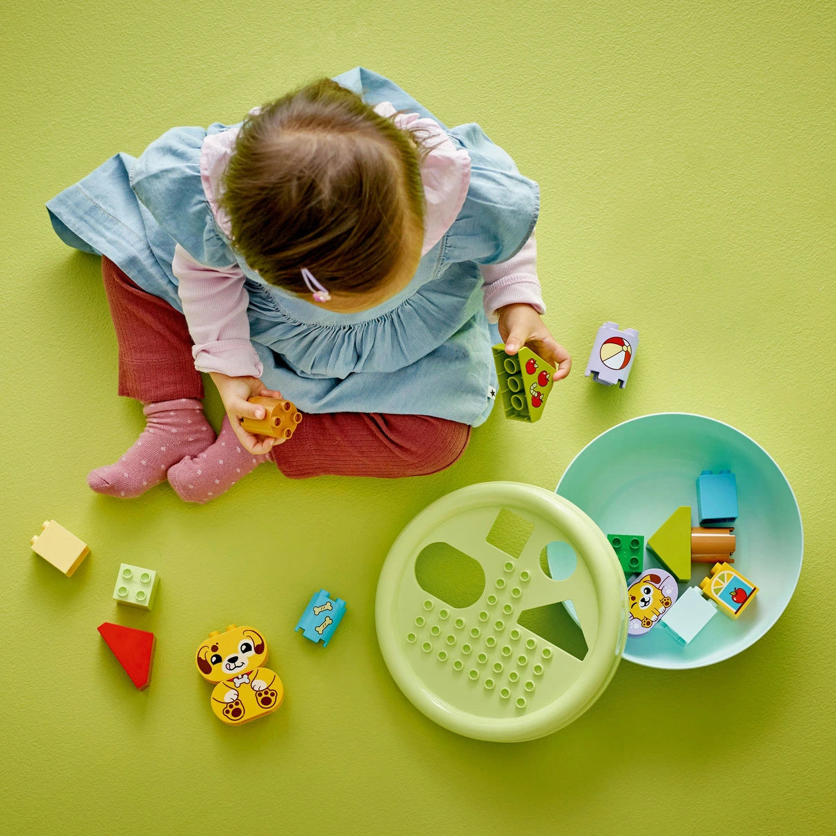 Kisgyermek ül világoszöld padlón és LEGO DUPLO építőkockákkal játszik; színes, állatmintás és formaberakós elemek veszik körül, a játék egy zöld fedelű tárolóedényből származik.
