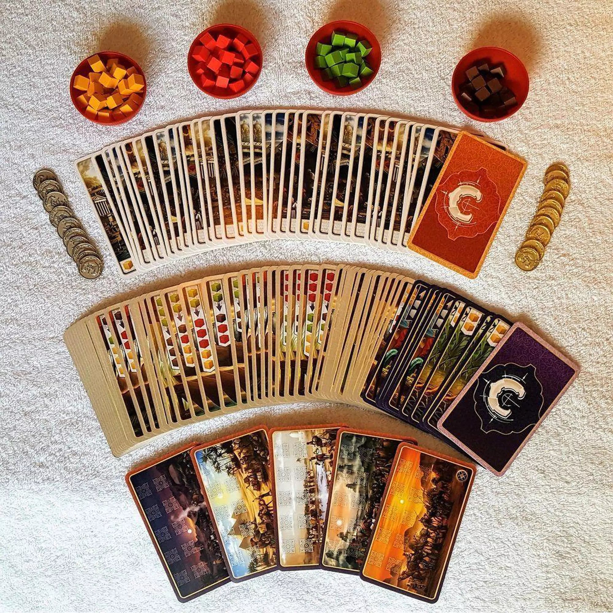 A fűszerkereskedelem társasjáték kártyái és nyersanyagok, köztük színes fa jelölők és pénzérmék, rendezve a játék előkészítéséhez.