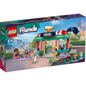 LEGO Friends 41728 Heartlake belvárosi büfé – hivatalos dobozkép, amely bemutatja az éttermet városi környezetben. A dobozon láthatóak a karakterek: Aliya, Liann és Charli, valamint az ikonikus szendvics a tetőn. A bal felső sarokban a korosztályi ajánlás (6+), alul a szereplők portréi.