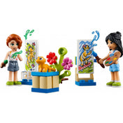 LEGO Friends 41739 Liann szobája – festőművészeti jelenet: Liann és Autumn minifigurák festőállvány előtt dolgoznak. Mindkét karakter élénk színű ruhákban látható, a festmények vidám állatokat ábrázolnak. A középen elhelyezett virágos dísz és sárga gyík teszi még játékosabbá a jelenetet.