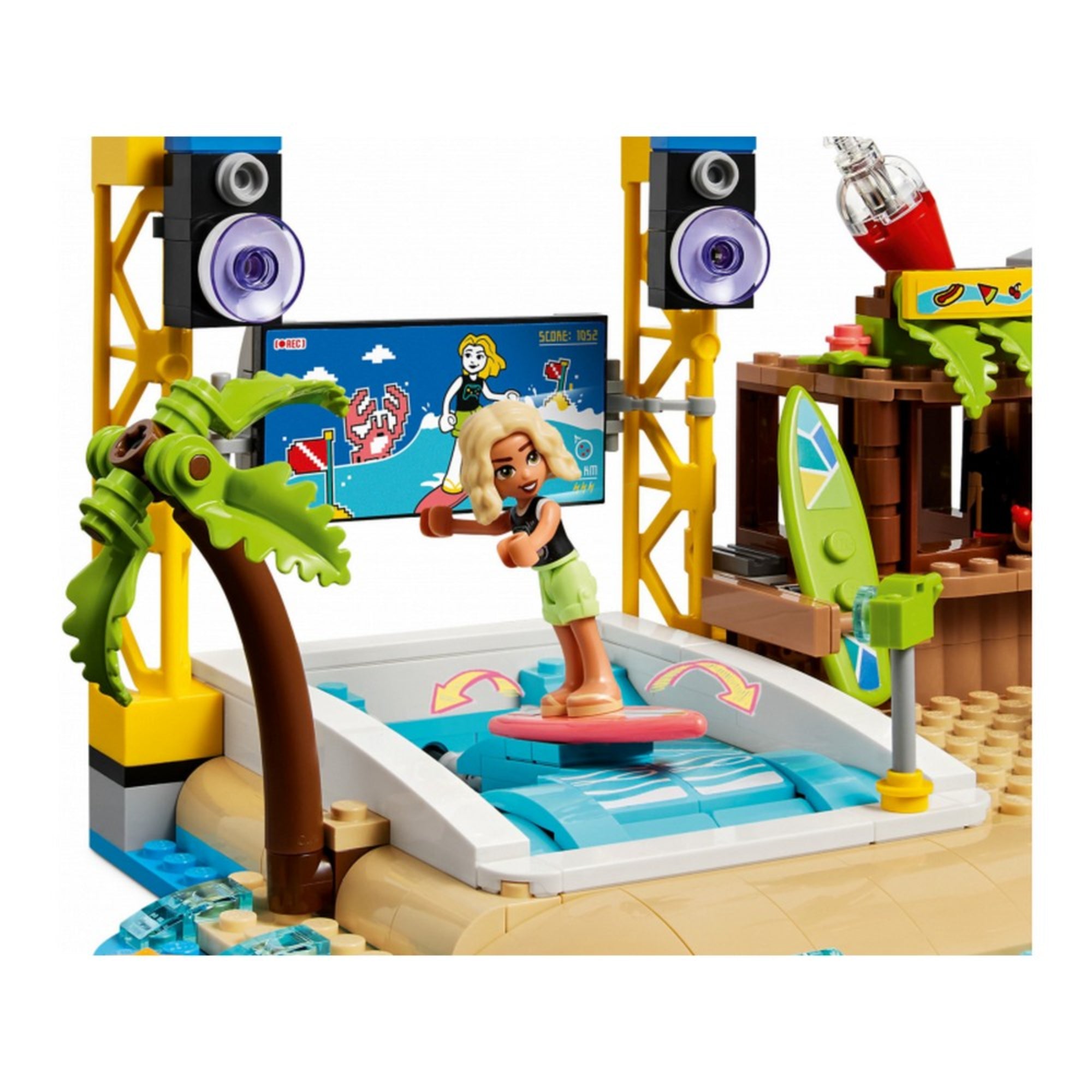 Egy szőke LEGO Friends figura szörfözik egy mozgó hullámszimulátoron, háttérben játékos tengerparti díszlet, pálmafával és hangszórókkal – LEGO Friends Tengerparti vidámpark (41737).