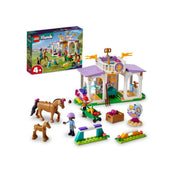 A LEGO Friends Új lovasiskola (41746) építőjáték doboza és a teljes készlet felépítve, ló figurákkal, karakterekkel, akadályokkal és a lovasiskola épületével egy vidám, zöld mezőn.