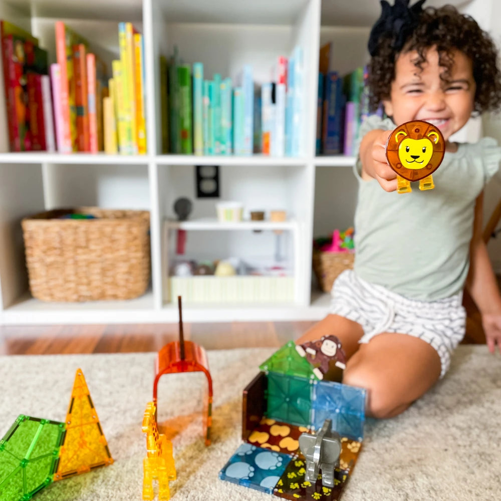 Kisgyermek mosolyogva mutat egy oroszlánt ábrázoló játékfigurát a kamera felé; előtte színes, áttetsző Magna-Tiles építőelemekből épített állatkertes jelenet látható, a háttérben könyvespolccal és fonott tárolókkal.