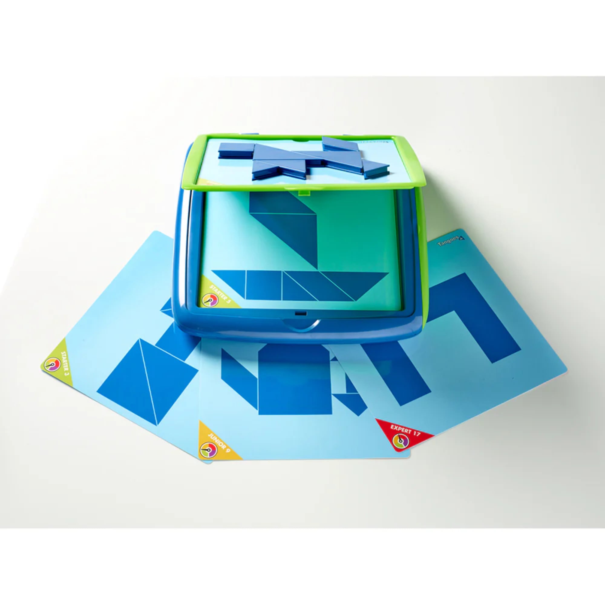 A képen a Smart Games Tangoes Junior logikai játék használat közben látható. A középen elhelyezkedő mágneses játéktáblán geometriai formákból álló kirakós feladat látható, amelyet a gyerekeknek kell megoldaniuk a hozzá tartozó kék tangram elemek segítségével. A tábla alatt több kihíváskártya található, különböző nehézségi szintekkel, például „Junior 9” és „Expert 17”. A színes sarokjelölések segítenek megkülönböztetni a kártyák nehézségi szintjét.