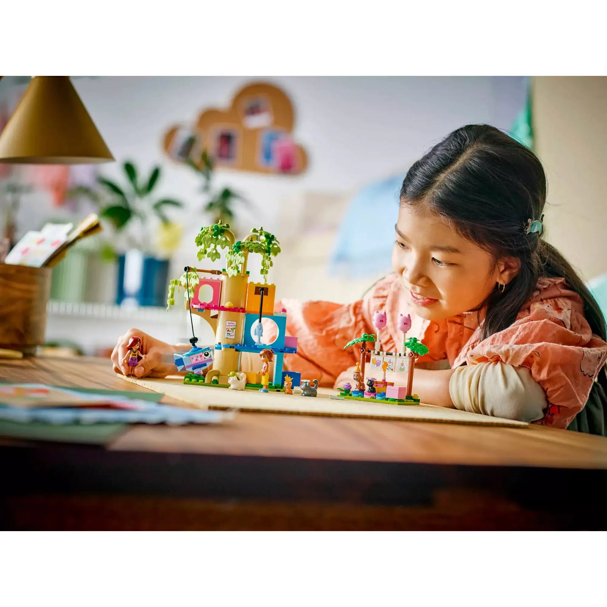 Egy kislány asztalnál játszik a LEGO Friends Macskaszülinapi buli (42666) készlettel, miközben a cicák és figurák egy születésnapi bulit rendeznek.