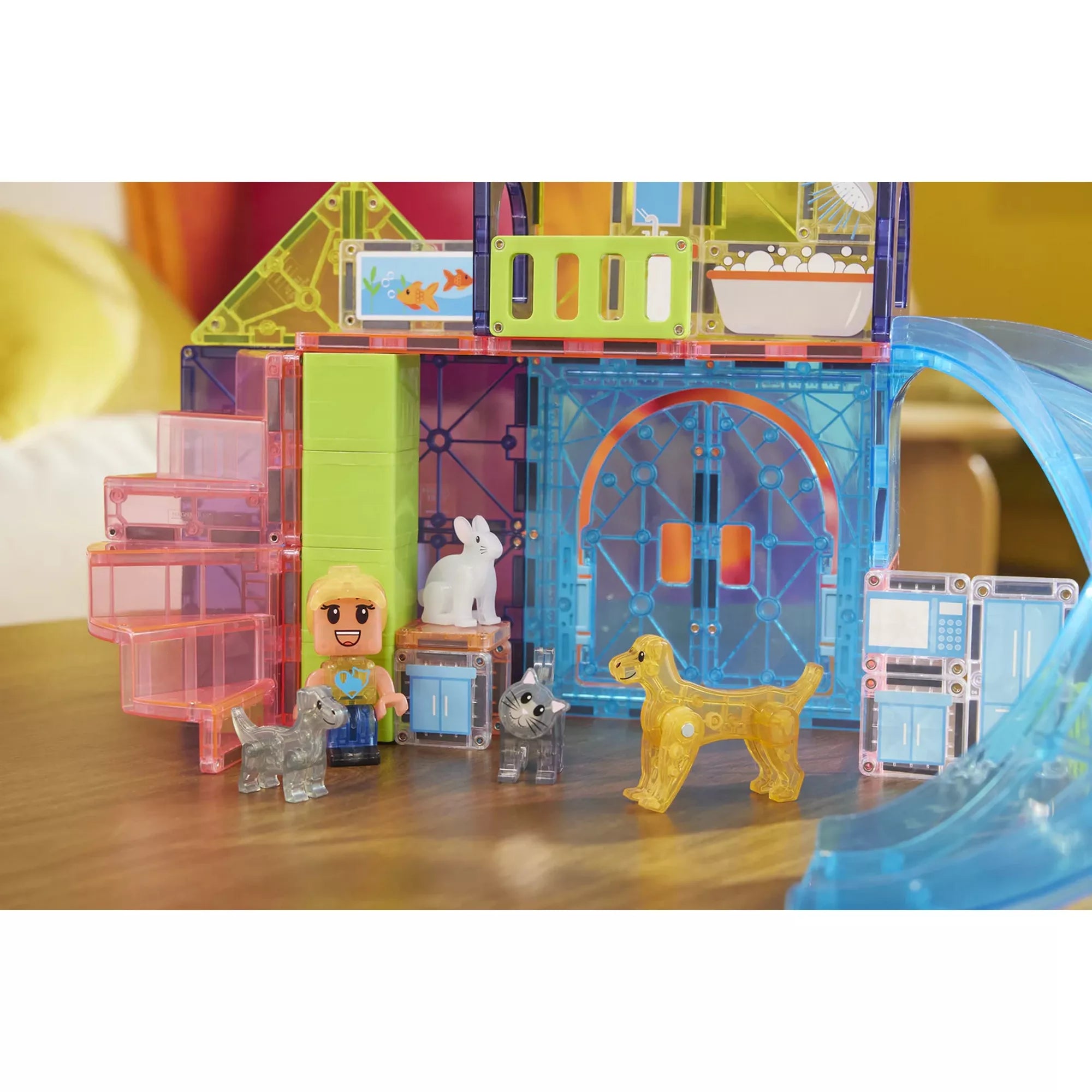 A MAGNA-TILES Pet Playhouse felépített játéka asztalon: a gondozófigura, a kutya, a cica és a nyuszi együtt játszanak a színes, áttetsző építőelemekből álló házban. A jelenet a gyerekek fantáziadús szerepjátékát mutatja be.