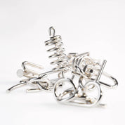 A 10 Metal Puzzle Set szétválasztott fémdarabjai fehér háttéren, különféle alakzatokkal: spirálrugó, szöges szerű elemek, hurkok és kampók, mind fényes ezüstös bevonattal, rendezetlenül egymásra helyezve.