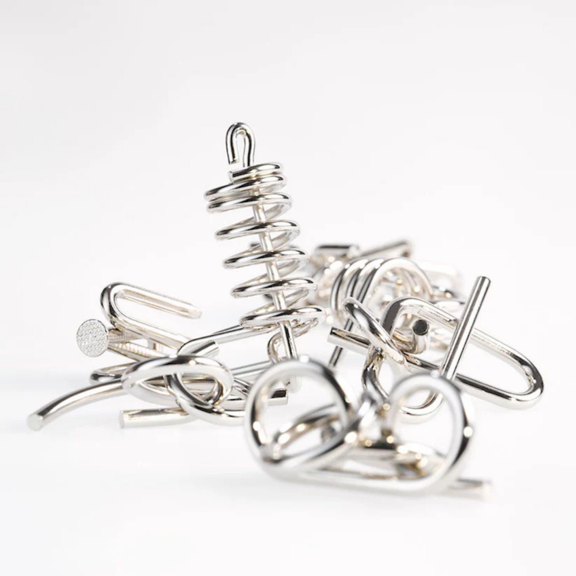 A 10 Metal Puzzle Set szétválasztott fémdarabjai fehér háttéren, különféle alakzatokkal: spirálrugó, szöges szerű elemek, hurkok és kampók, mind fényes ezüstös bevonattal, rendezetlenül egymásra helyezve.