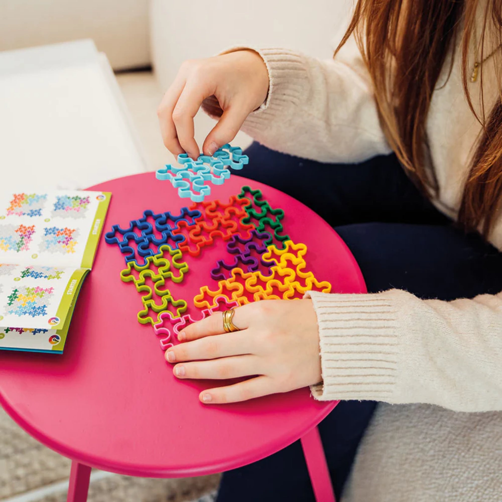 Az IQ Matrix logikai játék használat közben, ahol egy személy épp egy kék puzzle-darabot illeszt be a többi színes elem közé egy rózsaszín asztalon, mellette a kihívásokat tartalmazó füzet.