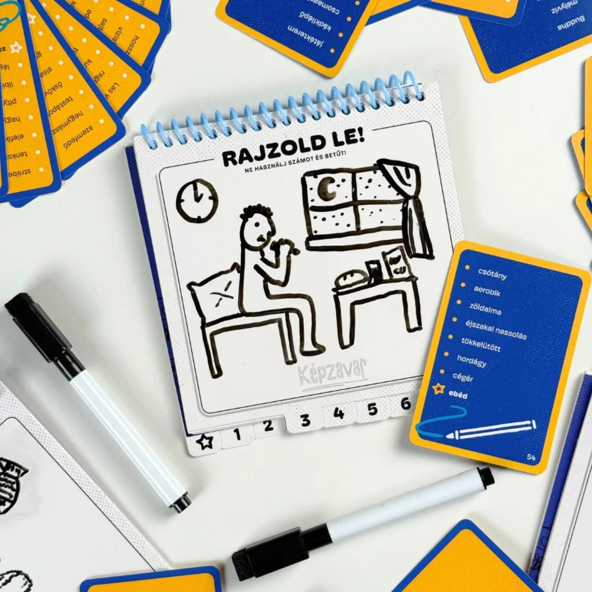 Képzavar társasjáték feladvány rajzfüzetben, egyszerű rajzzal egy esti jelenetről, mellette kártya és filctollak a játékhoz.