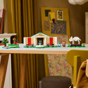 A LEGO 77056 készlet elemei egy modern faasztalon kiállítva. Középen a múzeum, két oldalán a jelenetépítő elemek – fa, víz, szökőkút és dinoszaurusz csontváz –, a háttérben lakásbelső és dekorációk láthatók.