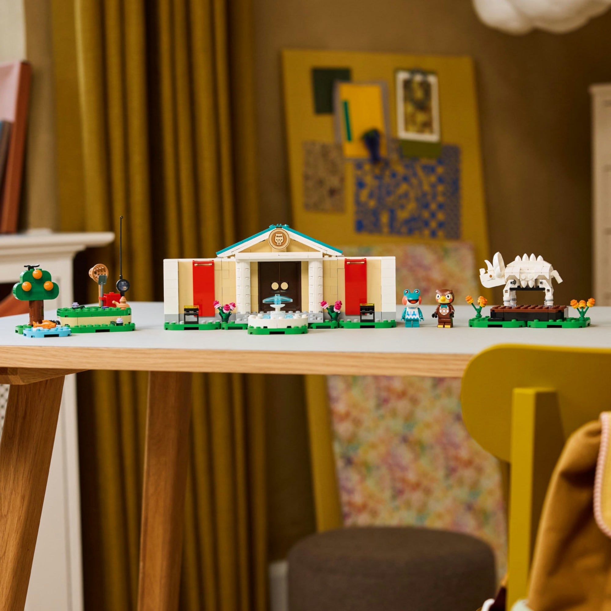 A LEGO 77056 készlet elemei egy modern faasztalon kiállítva. Középen a múzeum, két oldalán a jelenetépítő elemek – fa, víz, szökőkút és dinoszaurusz csontváz –, a háttérben lakásbelső és dekorációk láthatók.
