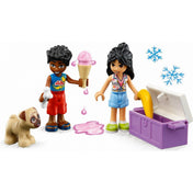 Közeli LEGO Friends jelenet a Homokfutó móka (41725) készletből: Zac piros rövidnadrágban rózsaszín fagyit tart, miközben egy aranyos mopsz kutyának csontot nyújt. Mellette Liann kék shortban és színes topban egy banánt vesz ki a lila hűtőládából. A háttérben két kék hógömb szimbólum emeli ki a hűtőláda jeges tartalmát. Vidám nyári pillanat LEGO minifigurákkal.