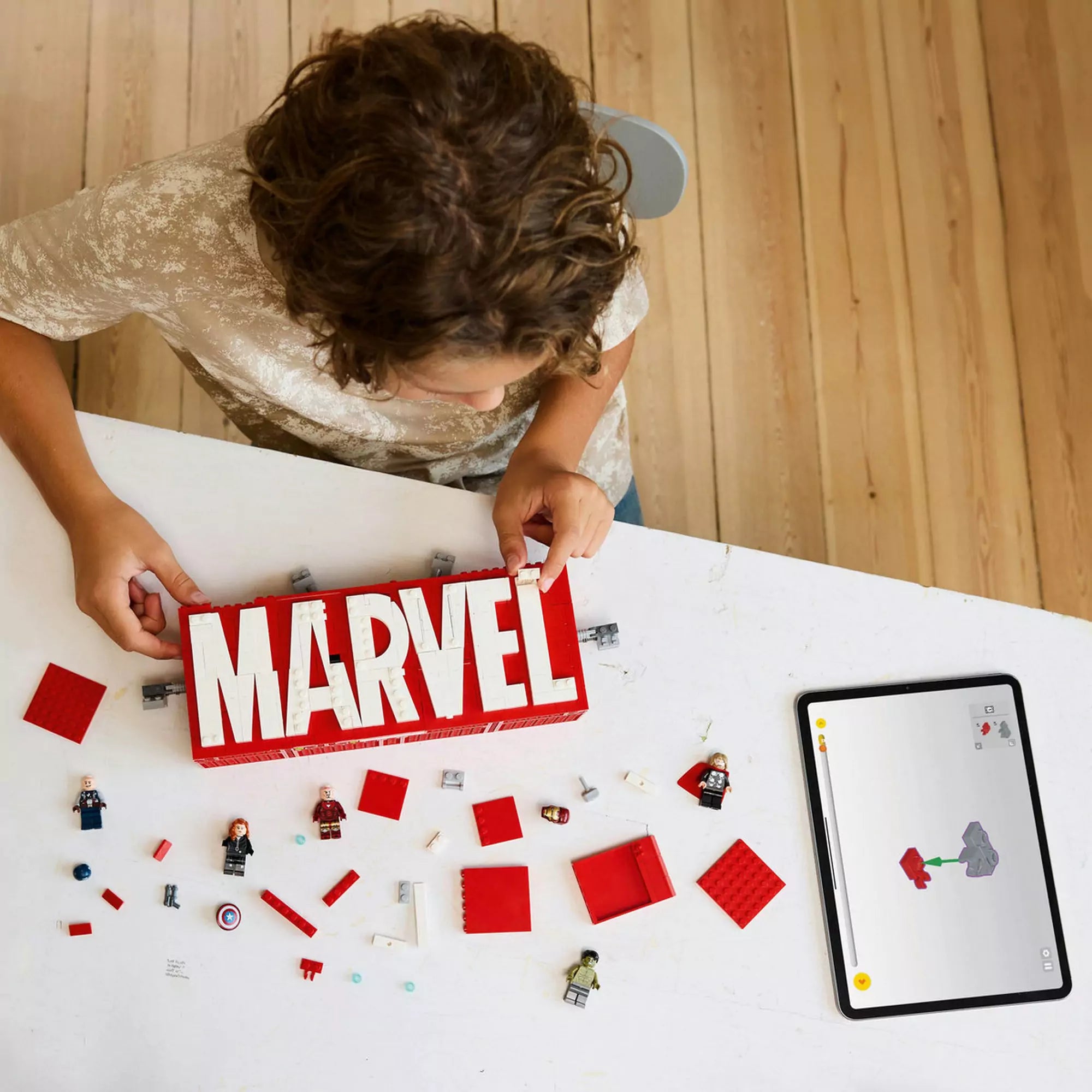 Gyermek építi a LEGO Marvel MARVEL logó és minifigurák (76313) készletet, minifigurák és kockák szétszórva az asztalon.