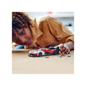 Egy fiatal lány játszik a teljesen elkészült LEGO Porsche 963 modellel egy asztalon, mellette néhány apró alkatrész látható.