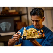 Egy férfi kézben tartja és építi a LEGO Star Wars Mos Espa fogatverseny diorámát (75380), miközben a háttérben egy sötét monitor és különböző dekorációk látszanak.
