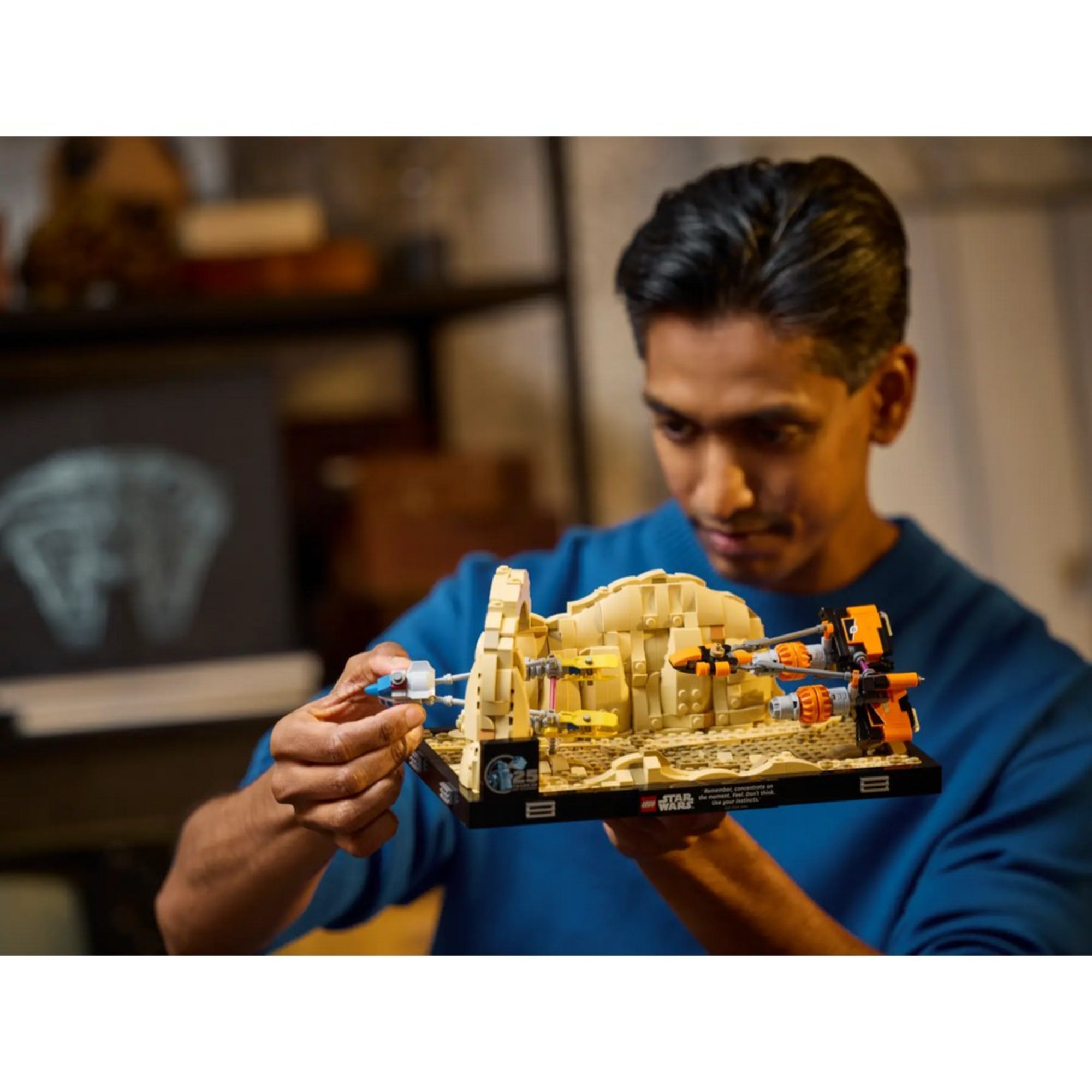 Egy férfi kézben tartja és építi a LEGO Star Wars Mos Espa fogatverseny diorámát (75380), miközben a háttérben egy sötét monitor és különböző dekorációk látszanak.
