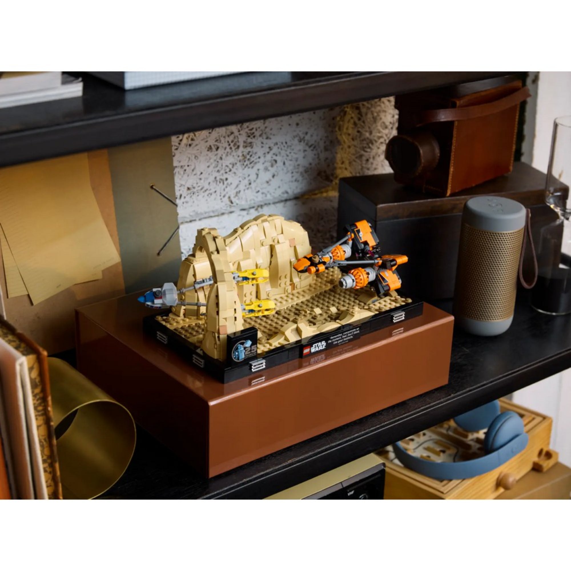 A LEGO Star Wars Mos Espa fogatverseny dioráma (75380) készlet egy fekete polcon elhelyezve, mellette modern hangszóró, fényképezőgép tok és más dísztárgyak láthatók.