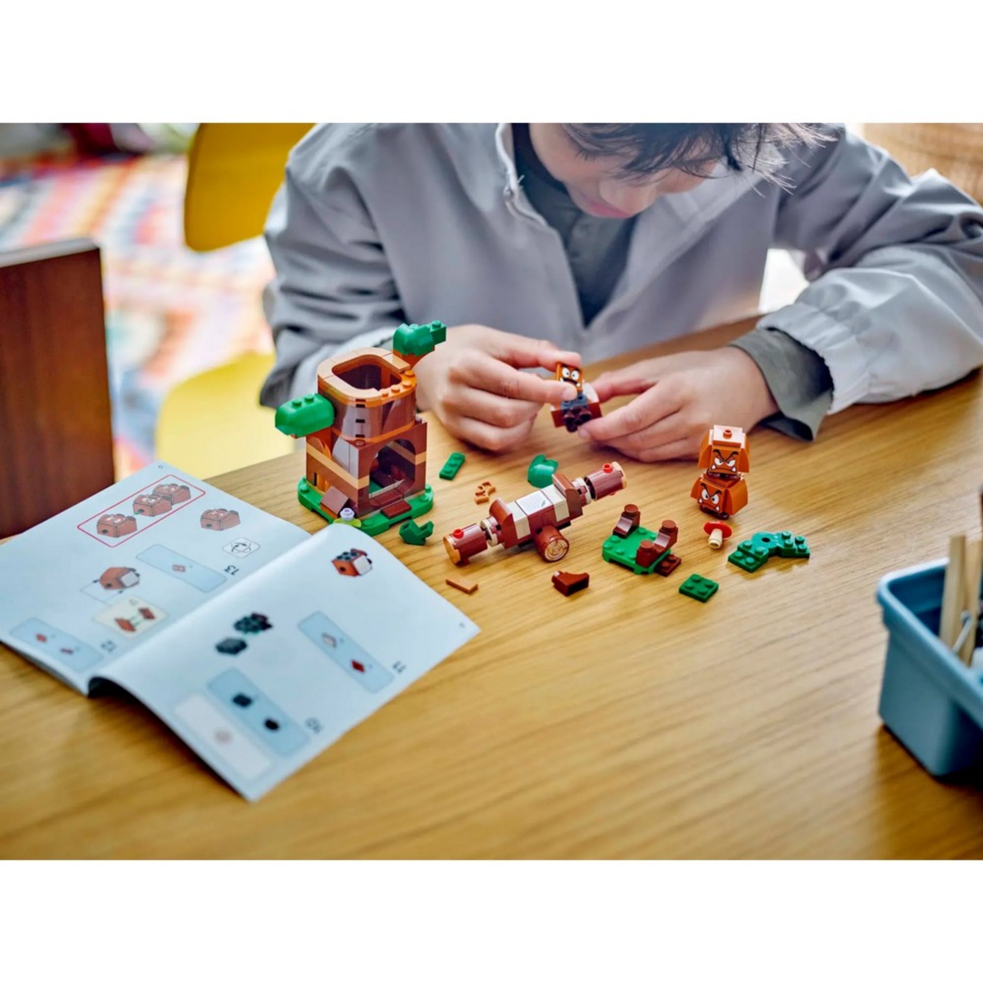Egy gyerek éppen összerakja a LEGO Super Mario Goomba játszótér (71433) készletet az asztalnál, az útmutató füzet segítségével.