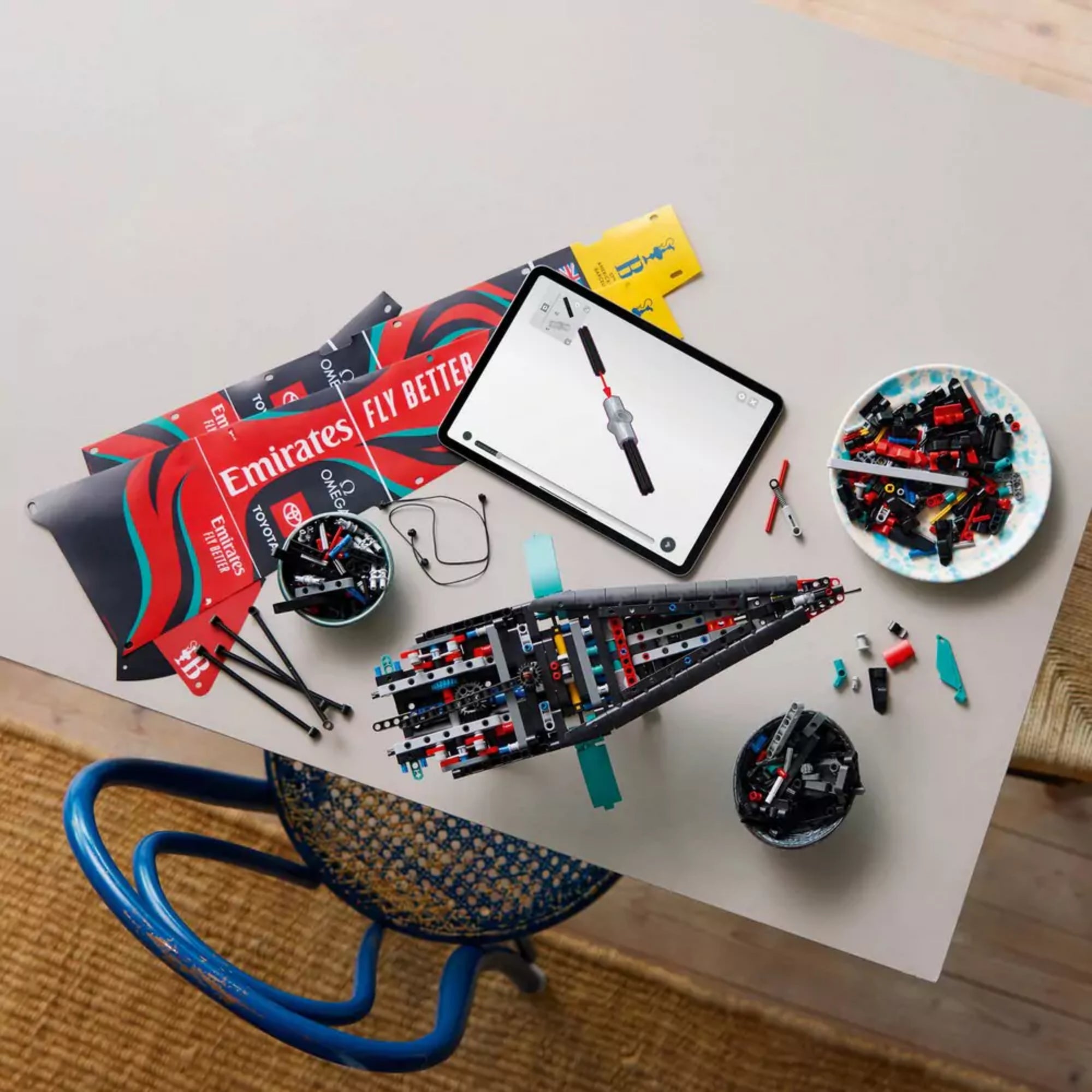 LEGO Technic Emirates Team New Zealand AC75 építés közben, egy asztalon elrendezett alkatrészekkel, tableten megjelenített digitális útmutatóval.