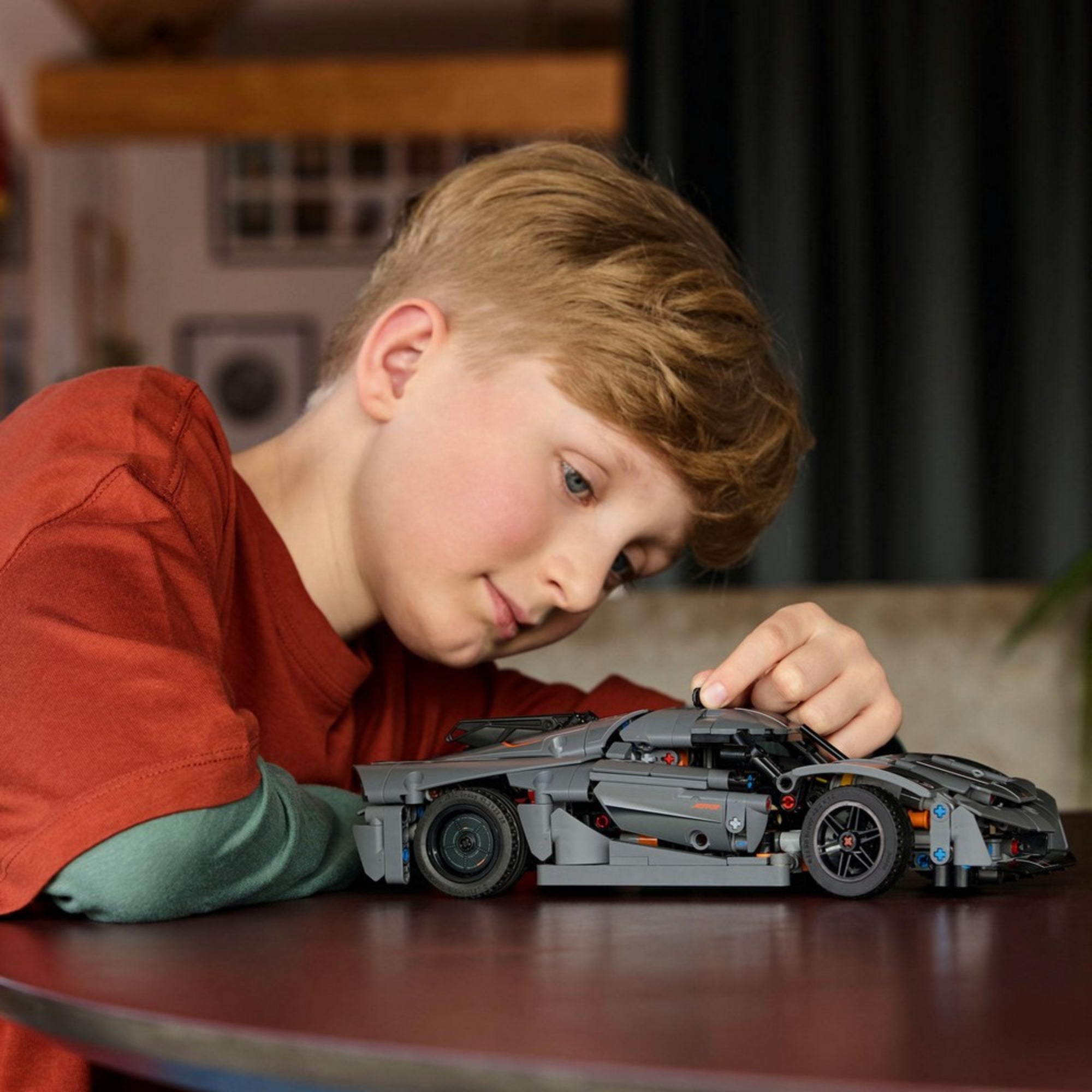 Egy fiú játszik a LEGO Technic Koenigsegg Jesko modellel, miközben mozgatja a jármű elemeit; a háttérben egy otthonos nappali látható.