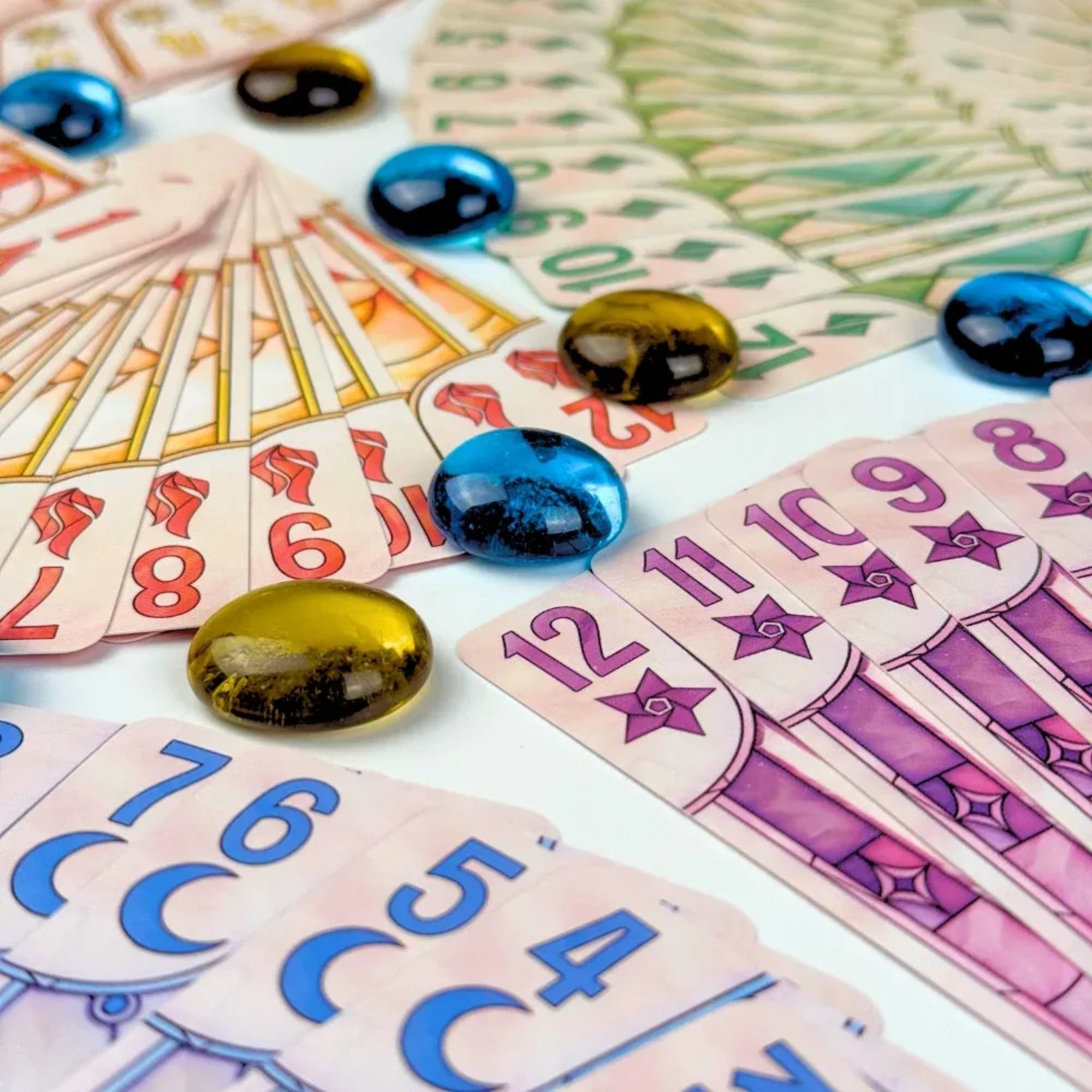 Közeli fotó a LUZ játékelemekről: számozott, színes kártyák legyezőszerűen elrendezve, rajtuk kék és arany üvegkavics jelölők.