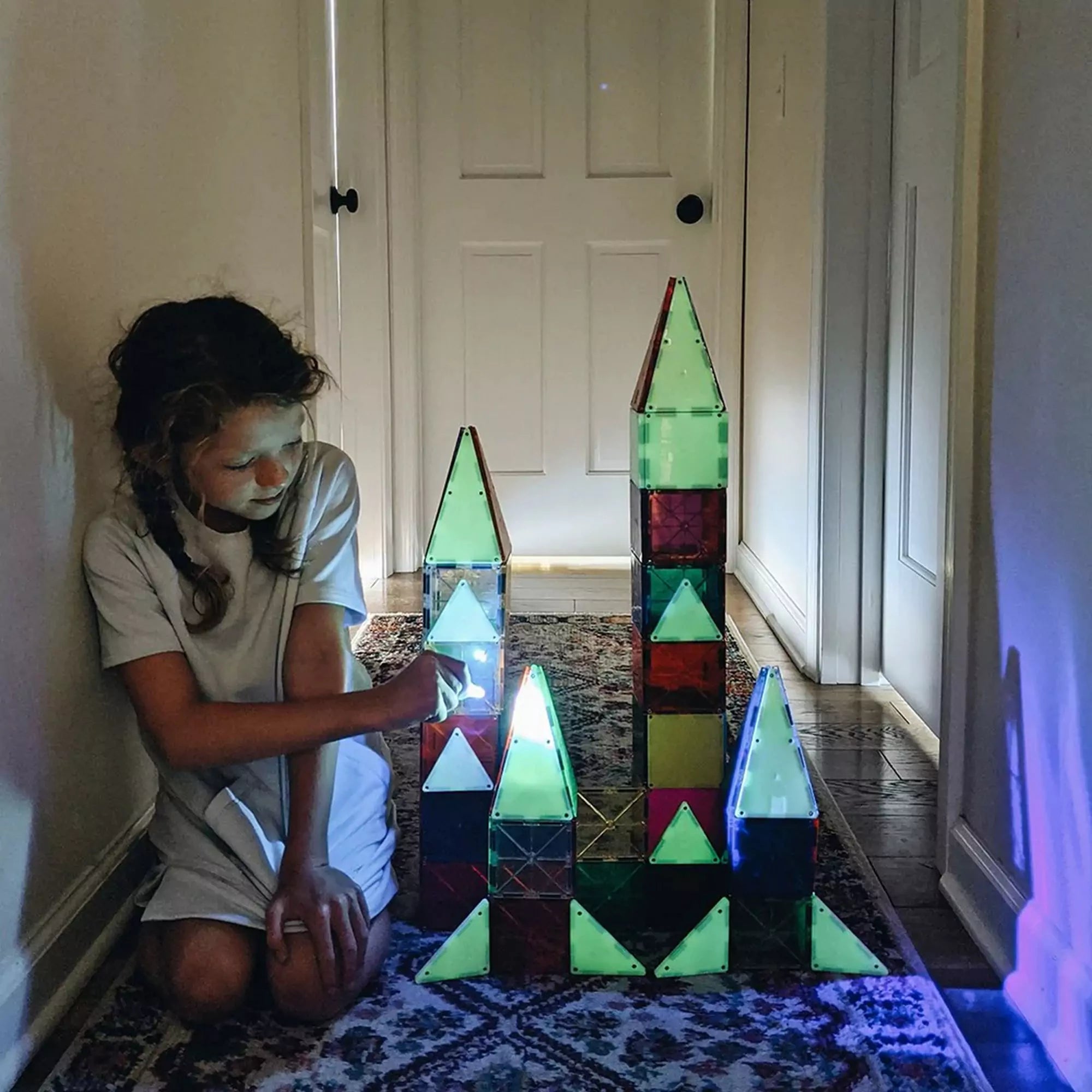 Egy kislány sötét folyosón játszik a Magna-Tiles Glow készlettel, egy világító kastélyt építve. Az elemek foszforeszkáló hatása lenyűgöző látványt nyújt.