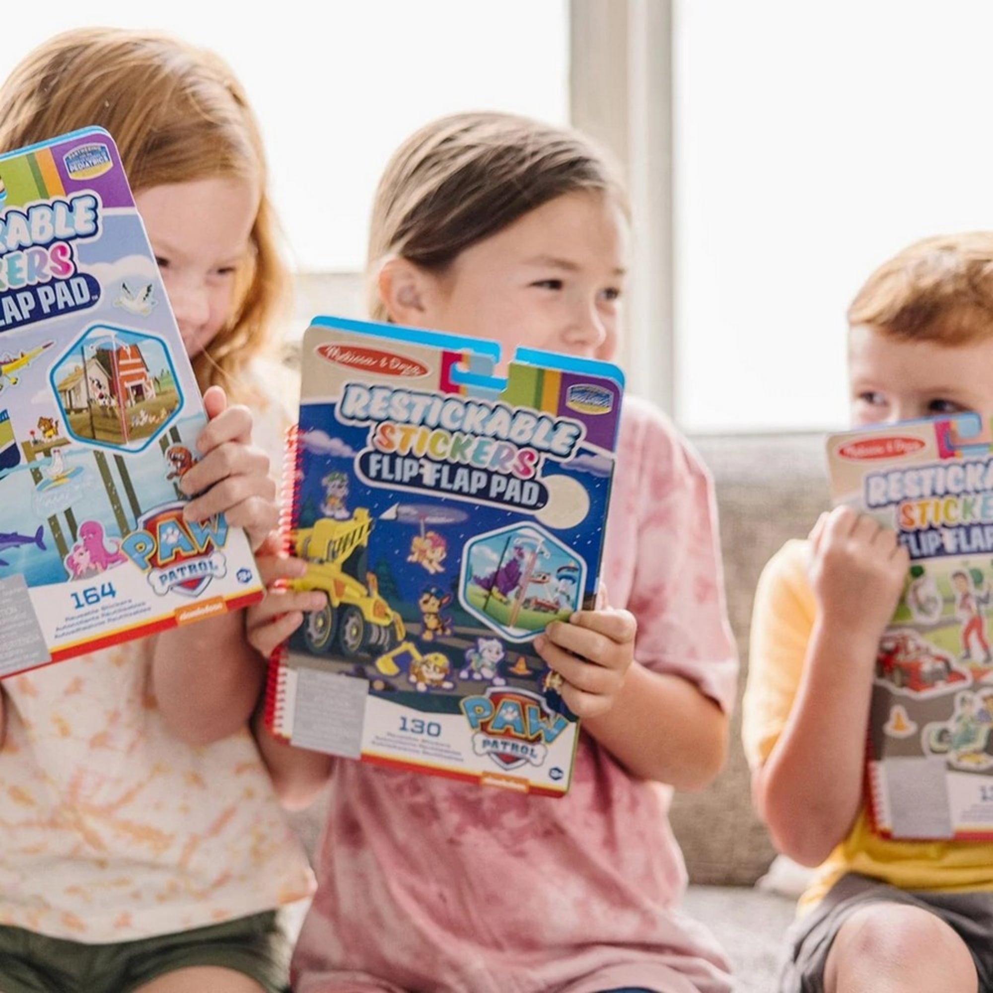 Mosolygó gyerekek kezükben tartják a Melissa & Doug Mancs őrjárat újrahasználható matricás füzeteket, különböző szetteket bemutatva.