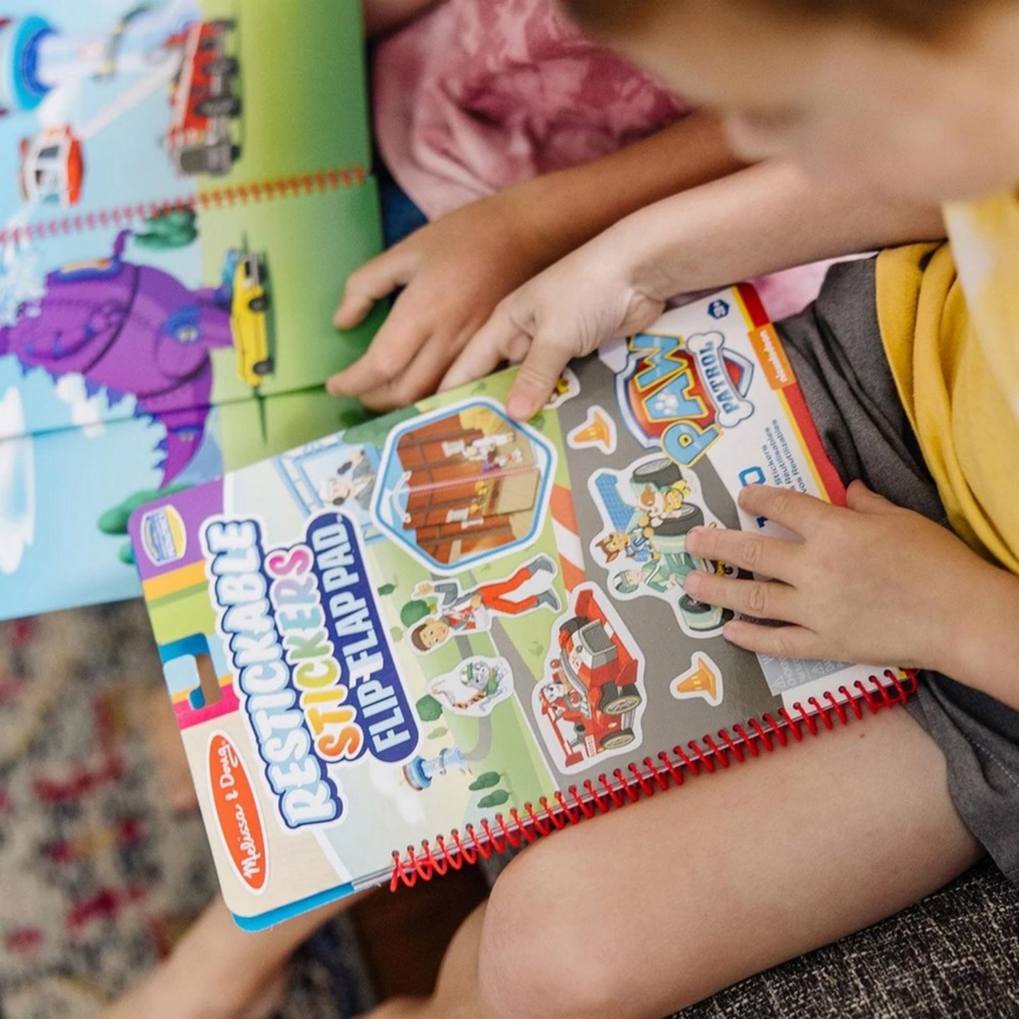 Két gyermek Mancs őrjárat matricás füzettel játszik, lapozzák a spirálos füzetet, és matricákat helyeznek el a vidám, színes jelenetekben.