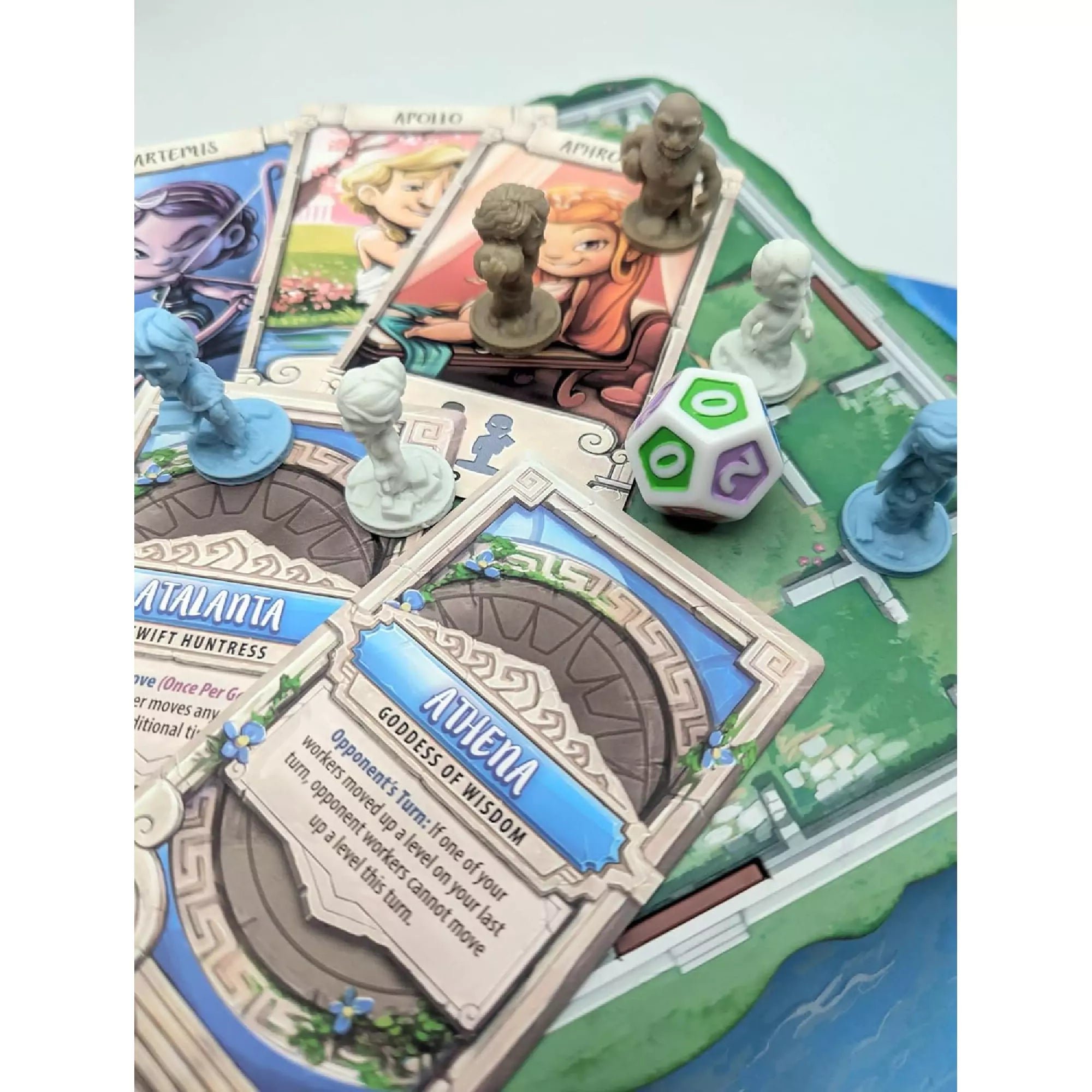 Santorini Second Edition társasjáték játékmenet közben, ahol kártyák, figurák és egy sokoldalú dobókocka látható a pályán.