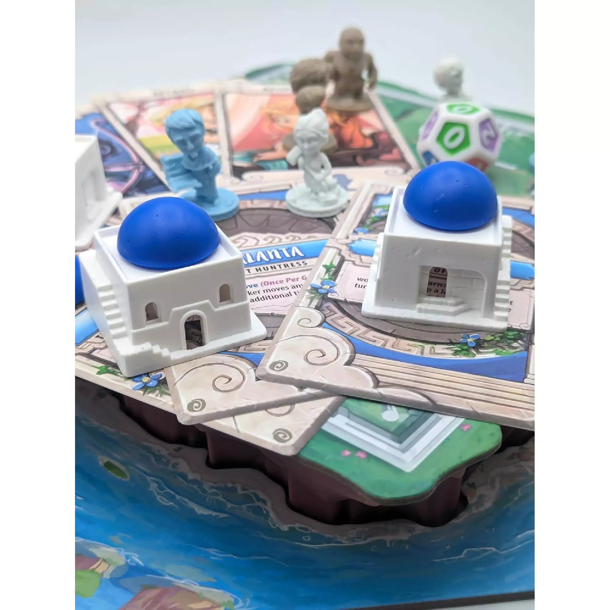 Santorini Second Edition társasjáték elemei közelről, fehér épületekkel, kék kupolákkal, istenkártyákkal és szoborszerű figurákkal.