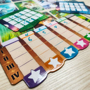 A Stella: Dixit Universe társasjáték pontozótábláinak közelképe, amelyeken színes csillagok és oszlopok láthatók, a játékosok pontszámainak nyomon követésére. A háttérben további játékkártyák és komponensek láthatók, amelyek a játék kreatív és vizuálisan lenyűgöző jellegét tükrözik.