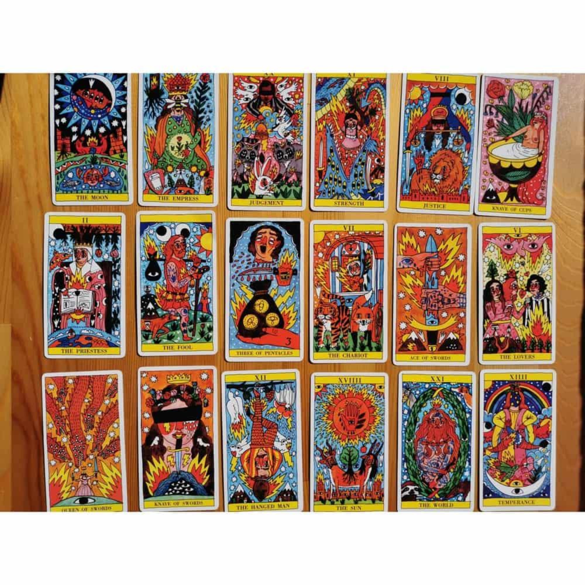 A képen a „Tarot del Fuego” kártyacsomag több lapja látható szétterítve, színes, kézzel rajzolt illusztrációkkal. Ricardo Cavolo stílusában készült grafikák vibráló színekben pompáznak: élénk vörös, sárga, narancs és kék tónusok dominálnak. A kártyákon mitikus és spirituális jelenetek jelennek meg, tűzmotívumokkal, szemekkel, szívekkel, napokkal és csillagokkal díszítve. A kártyák angol nyelvű felirattal vannak ellátva, pl. The Moon, The Fool, The Lovers.