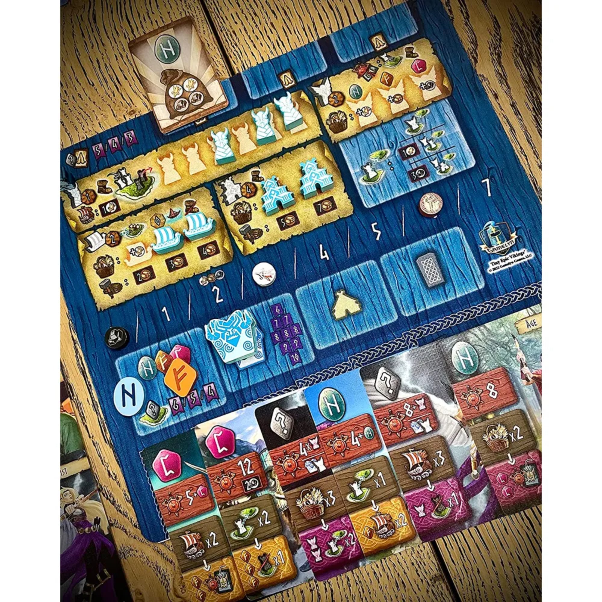 Közelkép a Tiny Epic Vikings játék egyik játéktáblájáról, ahol különféle épületek, hajók és viking ikonok láthatók, sokféle erőforrás-jelölővel.