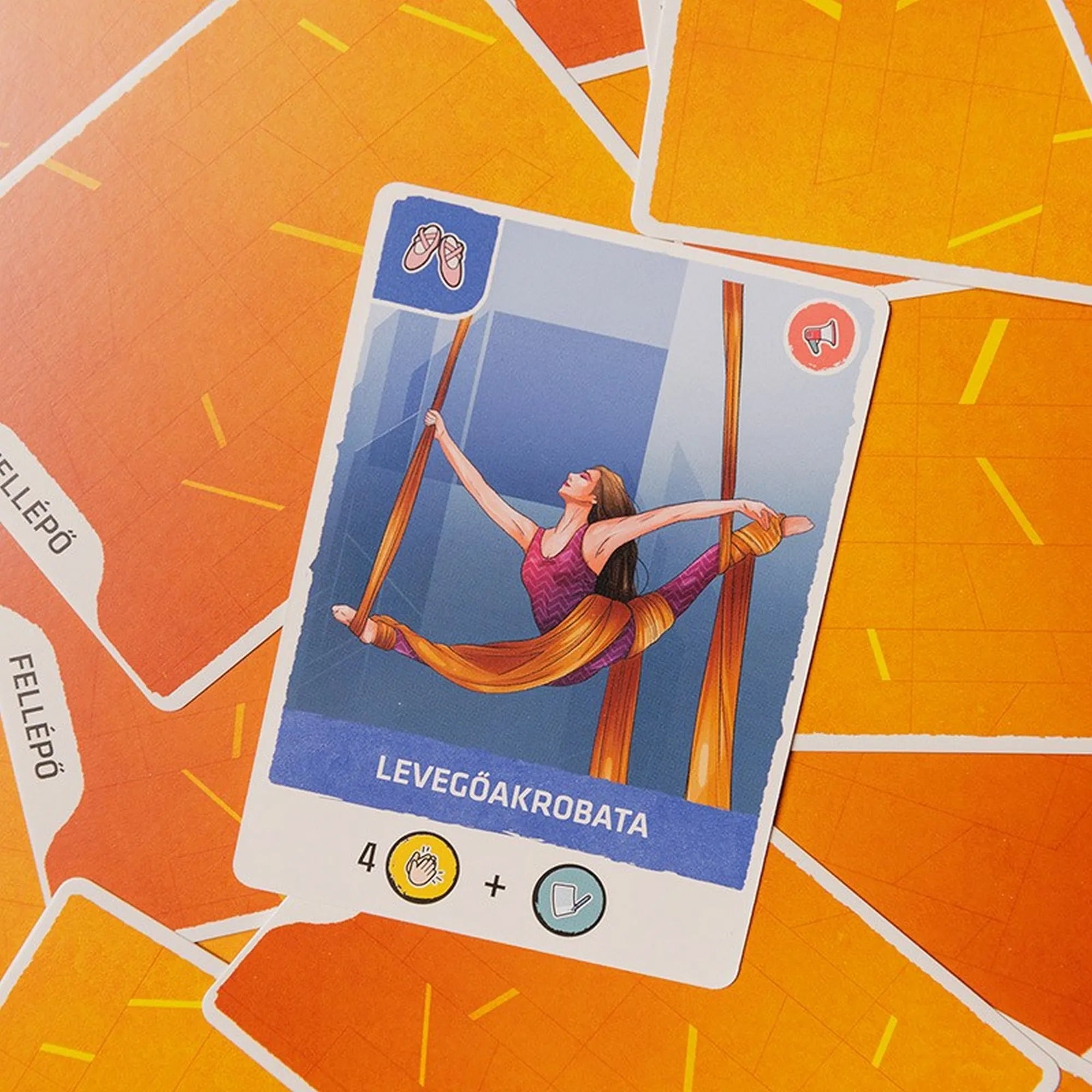 Az Évad társasjáték levegőakrobata kártyája közelről, narancssárga hátlapos kártyákon, a művész szövetfüggönyökön függeszkedve.