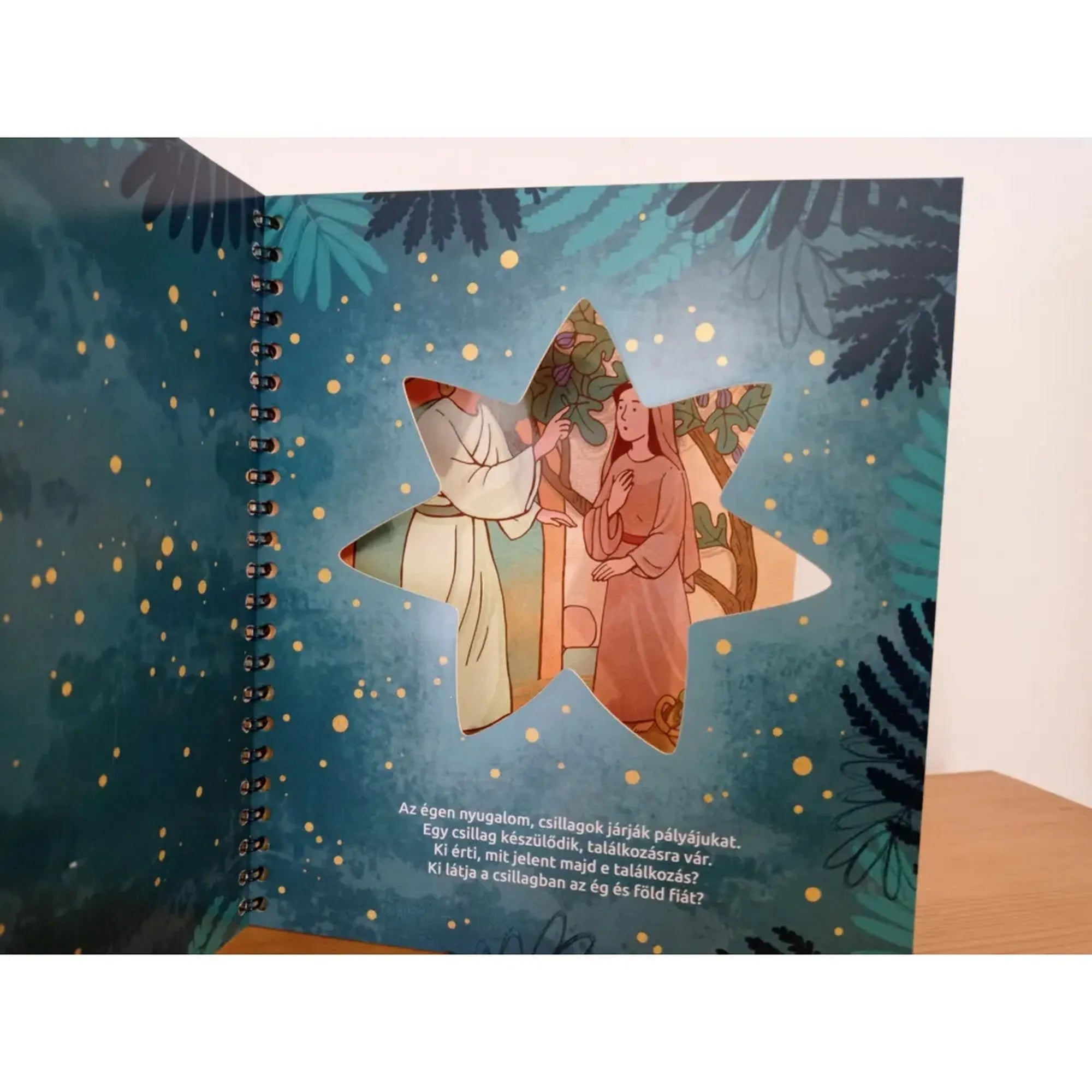 Az Ég és Föld Fia könyv oldala csillag alakú kivágással, amelyen át Mária és Gábriel találkozásának illusztrációja látszik.