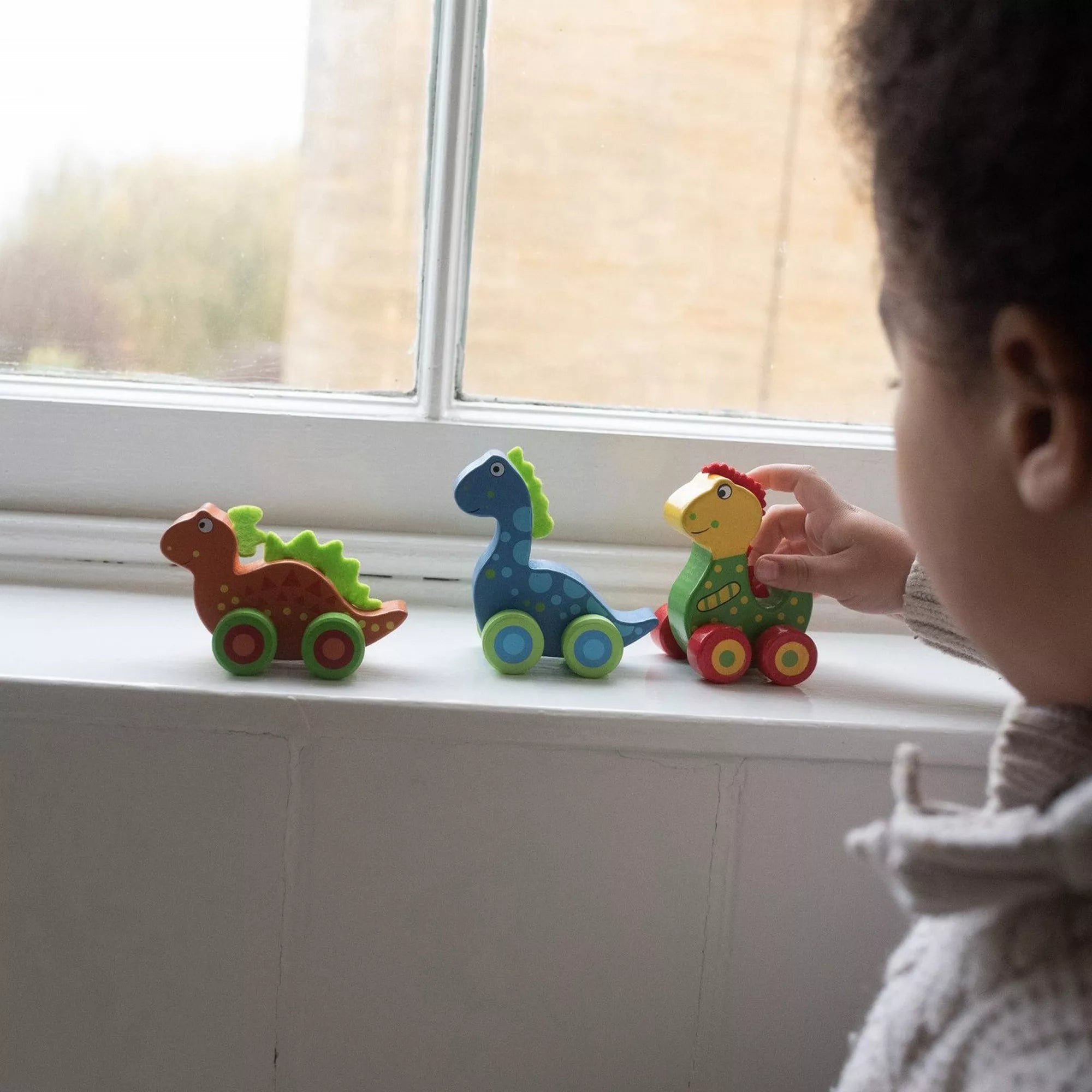 Egy kisgyermek az ablakpárkányon játszik az Orange Tree Toys színes, kerekes fa dinoszauruszaival – barna, kék és zöld játékfigurákkal.