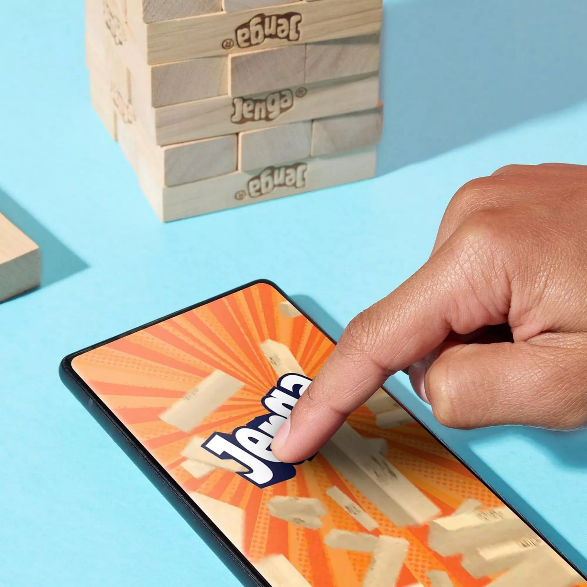 „Kéz egy okostelefon kijelzőjén a Jenga alkalmazás ikonra mutat, háttérben Jenga toronnyal, világoskék alapon.”