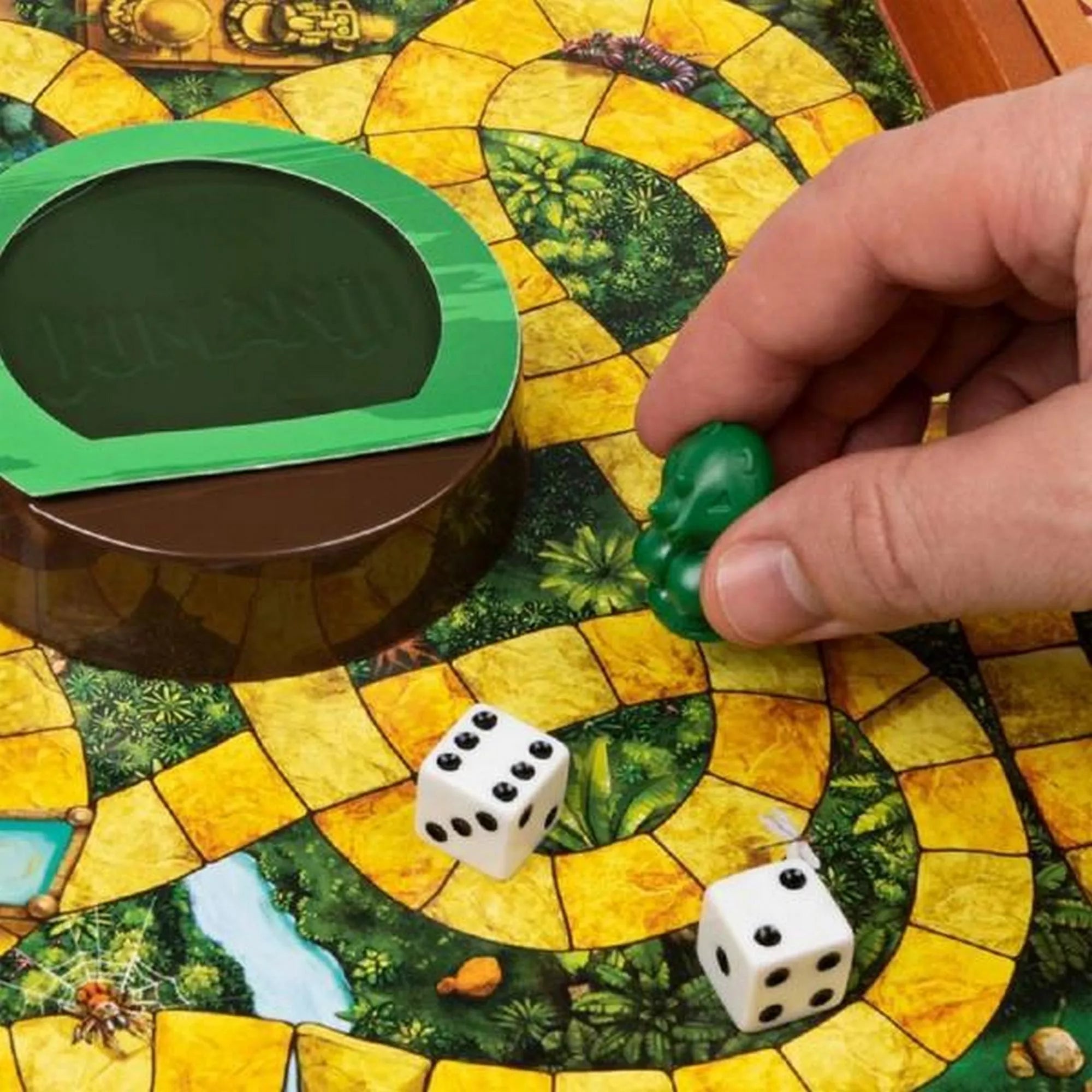 Közelkép a Jumanji társasjáték játéktáblájáról, amely dzsungel mintázatú, sárga ösvényekkel. Egy játékos keze zöld bábut helyez a pályára a dobókockák mellé, a középen található zöld Jumanji kupola mellett.