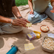 Két ember a szabadban, egy pokrócon ülve játszik a Legjobb barátok kártyajátékkal. A képen látható a nyitott doboz, sárga hátlapú kártyák és egy tál popcorn, amelyek baráti, oldott hangulatot teremtenek.
