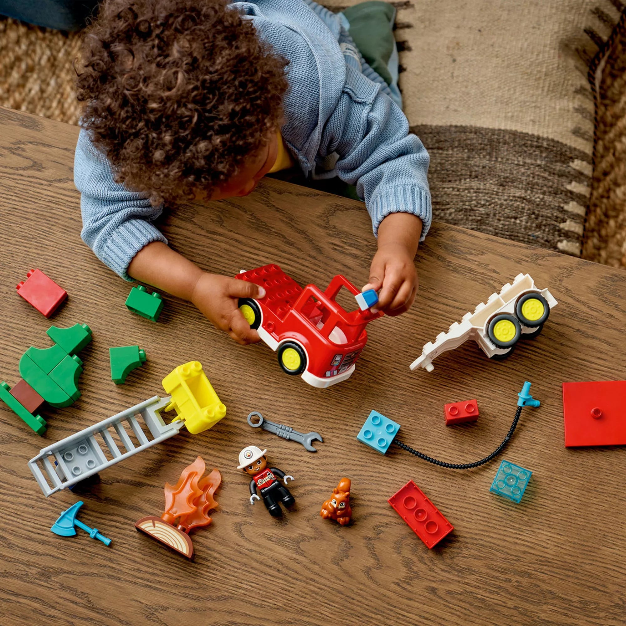 Kisgyermek a LEGO DUPLO 10473 Tűzoltóautó készlet elemeivel játszik, szétszedett járműrészekkel, létrával, láng elemmel, figurákkal és szerszámokkal a padlón.