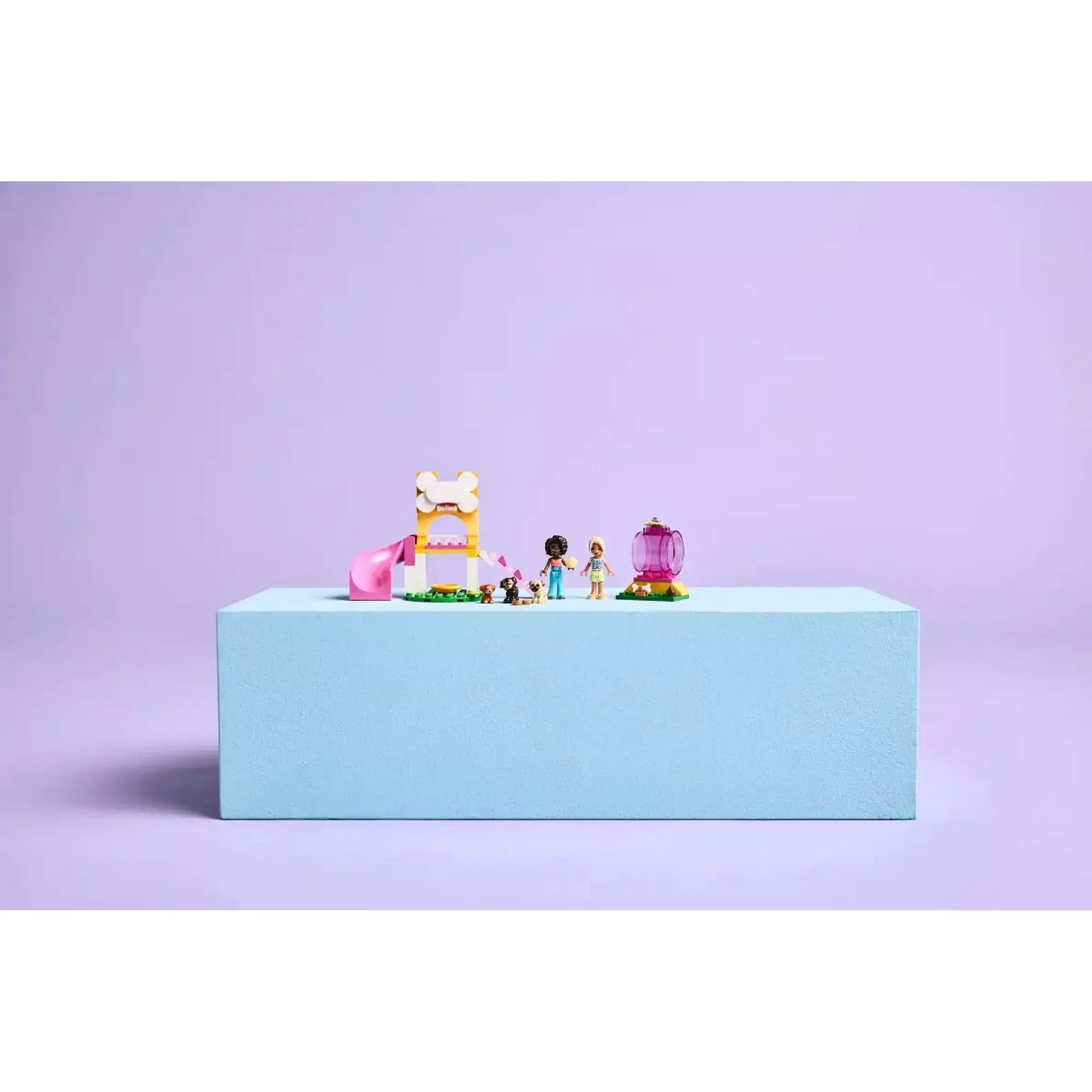 A LEGO Friends Puppy Playground (42665) készlet bemutatása világoslila háttéren, a készlet minden eleme külön elhelyezve egy világoskék emelvényen.