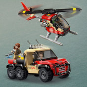 „LEGO Jurassic World (76976) járművek akcióban: a vörös-sárga helikopter a levegőben repül, alatta pedig a masszív, hatkerekű terepjáró halad előre, benne egy minifigurával. A jelenetben egy másik minifigura is látható, aki egy kék lövedékkel felszerelt kábítófegyvert tart, készen az expedícióra.
