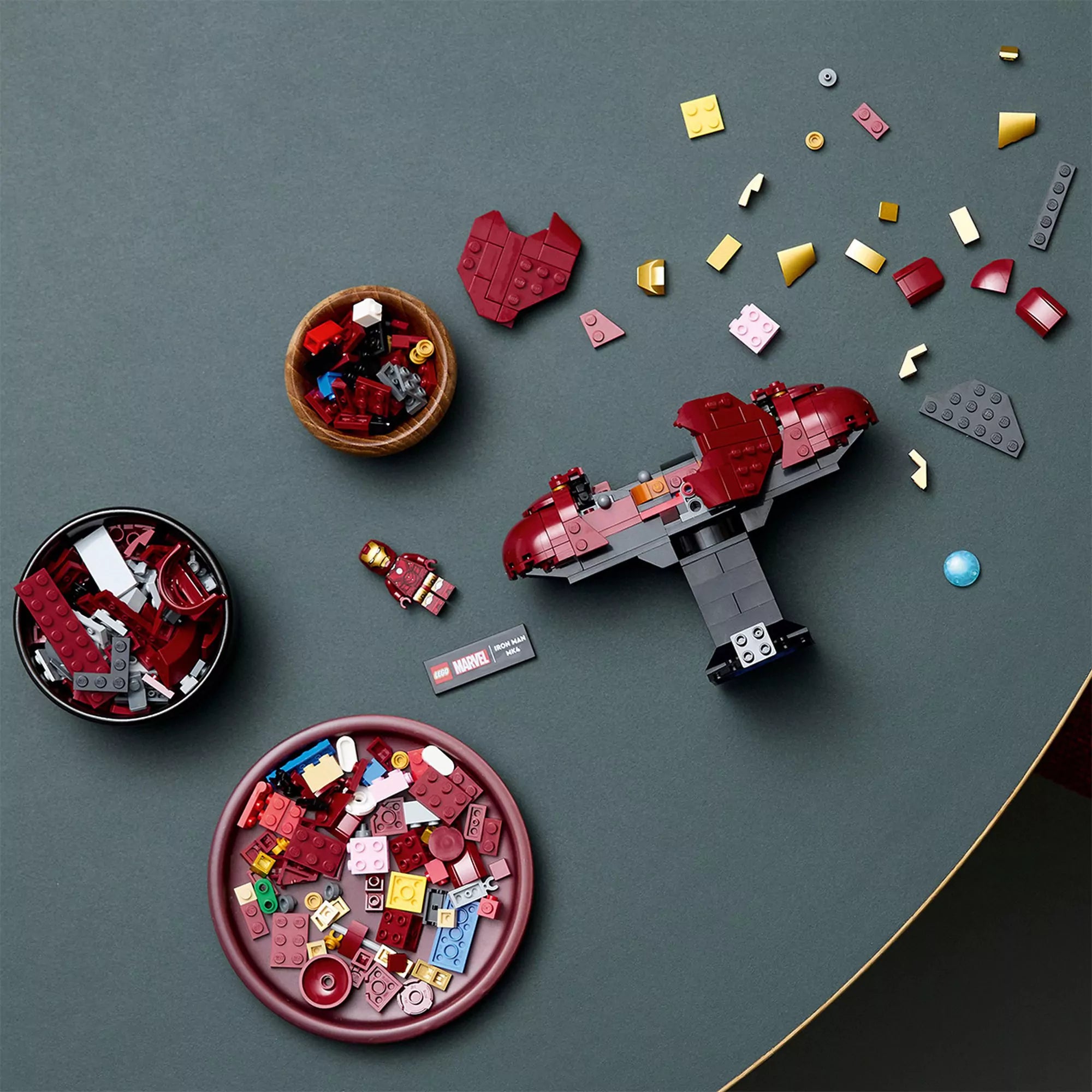 A LEGO Marvel Iron Man MK4 mellszobor (76327) félkész állapotban az asztalon, körülötte piros, arany és szürke LEGO elemek tálakban elhelyezve.