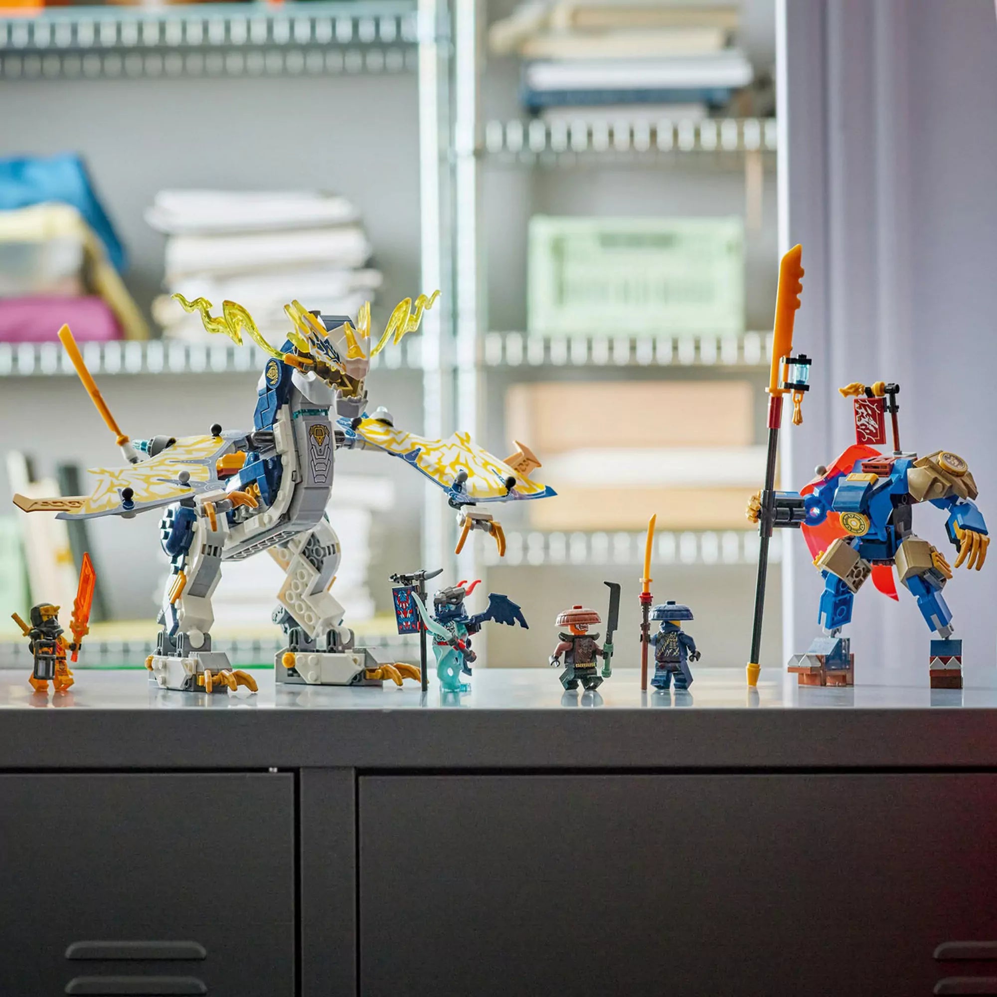 LEGO NINJAGO 71843 Rogue gépmechje és sárkánylovasa figurái és modelljei egy asztalon kiállítva, világos szobabelső előtt.