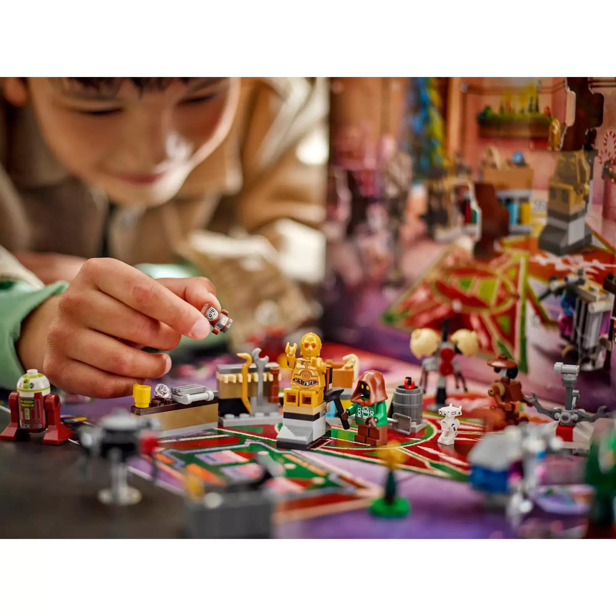 Gyerek kezében egy LEGO droidfigura, előtérben C-3PO, Jawa és különféle ünnepi miniépítmények a LEGO Star Wars Adventi naptár 2025 készletből.