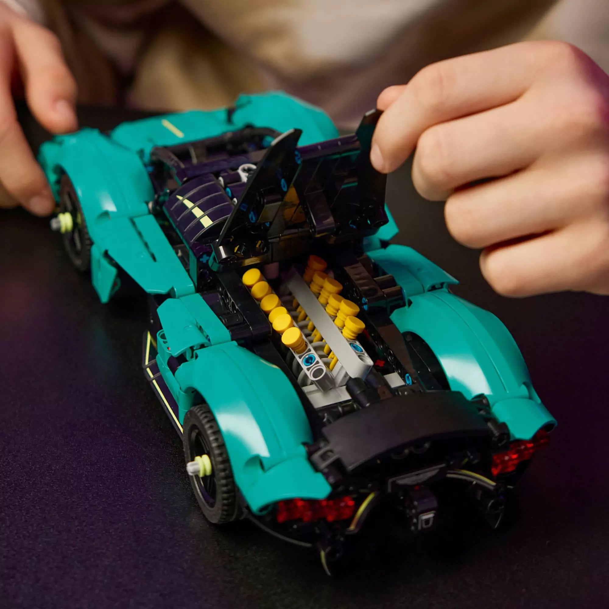  A LEGO Technic Aston Martin Valkyrie (42208) modell nyitott motorháztetője, alatta a sárga dugattyúkkal és részletes motorblokkal, egy gyermek kezében.