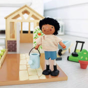 Mr. Forrester és a kutyája, Tender Leaf Toys TL8149 – Mr. Forrester a kertben áll egy öntözőkannával, háttérben egy üvegház és kerti szerszámok láthatók.