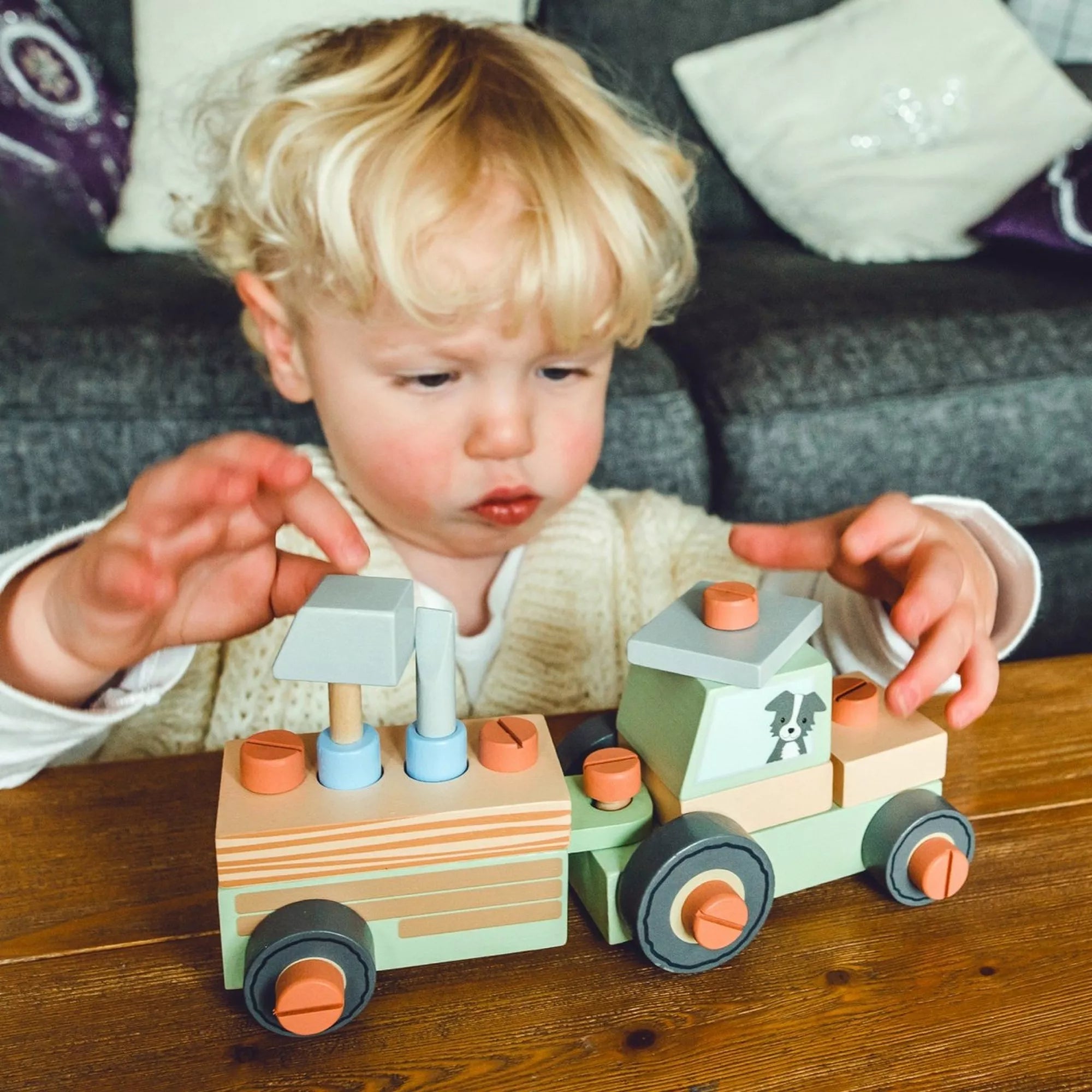 Egy kisgyermek játszik az Orange Tree Toys fa traktorral. A gyermek az asztalnál ül, miközben a traktor elemeit illeszti össze. A jelenet a játék kézzelfogható, tanulást segítő jellegét hangsúlyozza, ideális 1 éves kor felett.